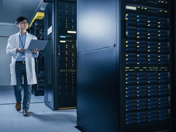 In Data Center: Male IT Technician Wearing White Coat Walking alongside Server Racks, Uses Laptop Computer to Run Maintenance Diagnostics Tools. He is Wearing Lab Coat and Working with Data.